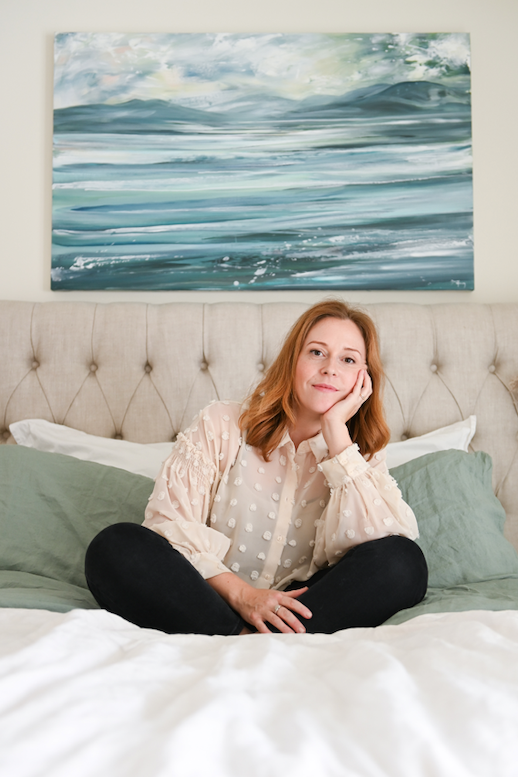 Canadian artist Dana Mooney sits cross legged on a bed with her green landscape painting hanging on the wall behind her above the tufted headboard.  